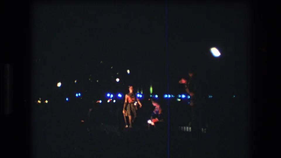 The 1977 Honolulu home-movie frame shows a dancer holding a staff-like prop beside another performer, with blue lights across the dark background.