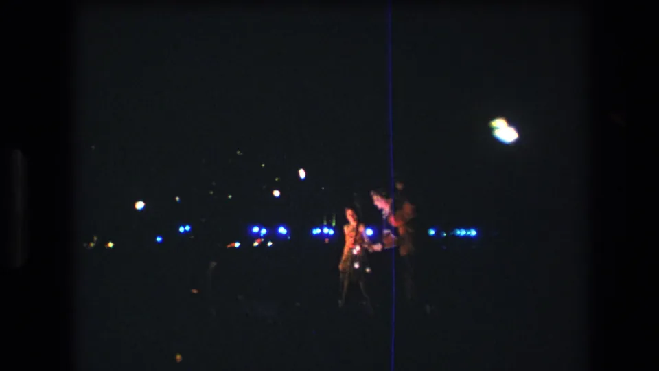 Two performers in traditional-style stage attire are visible in the 1977 Honolulu night footage, framed by blue background lights and the dark 8mm film image.