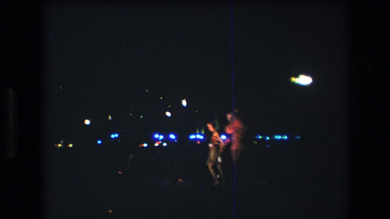 1977 Honolulu home-movie frame showing several people standing at night, with a brightly lit figure and scattered blue-white lights across the dark background.