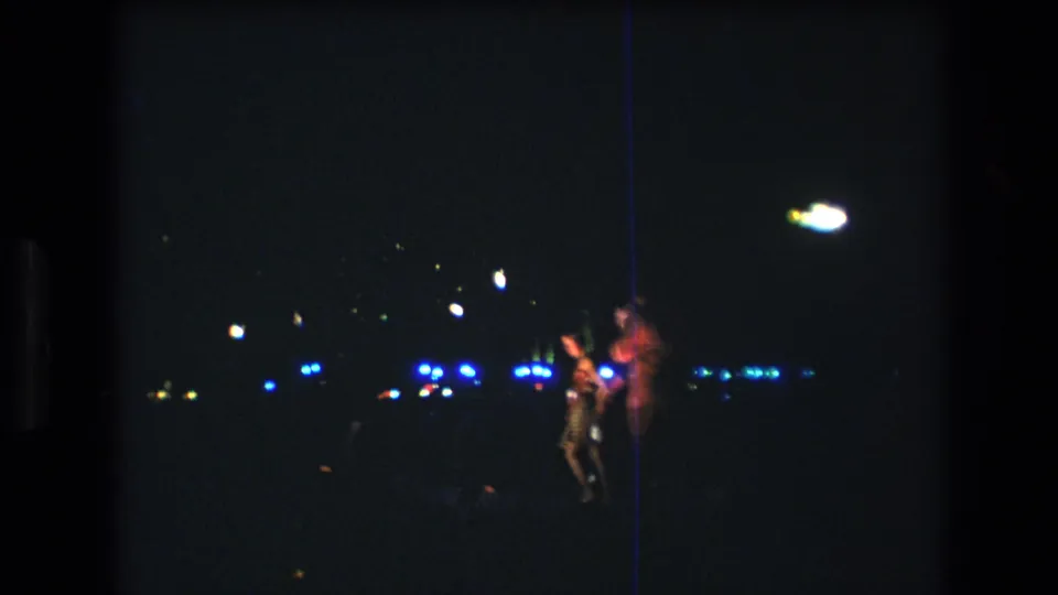 1977 Honolulu home-movie frame showing several people standing at night, with a brightly lit figure and scattered blue-white lights across the dark background.