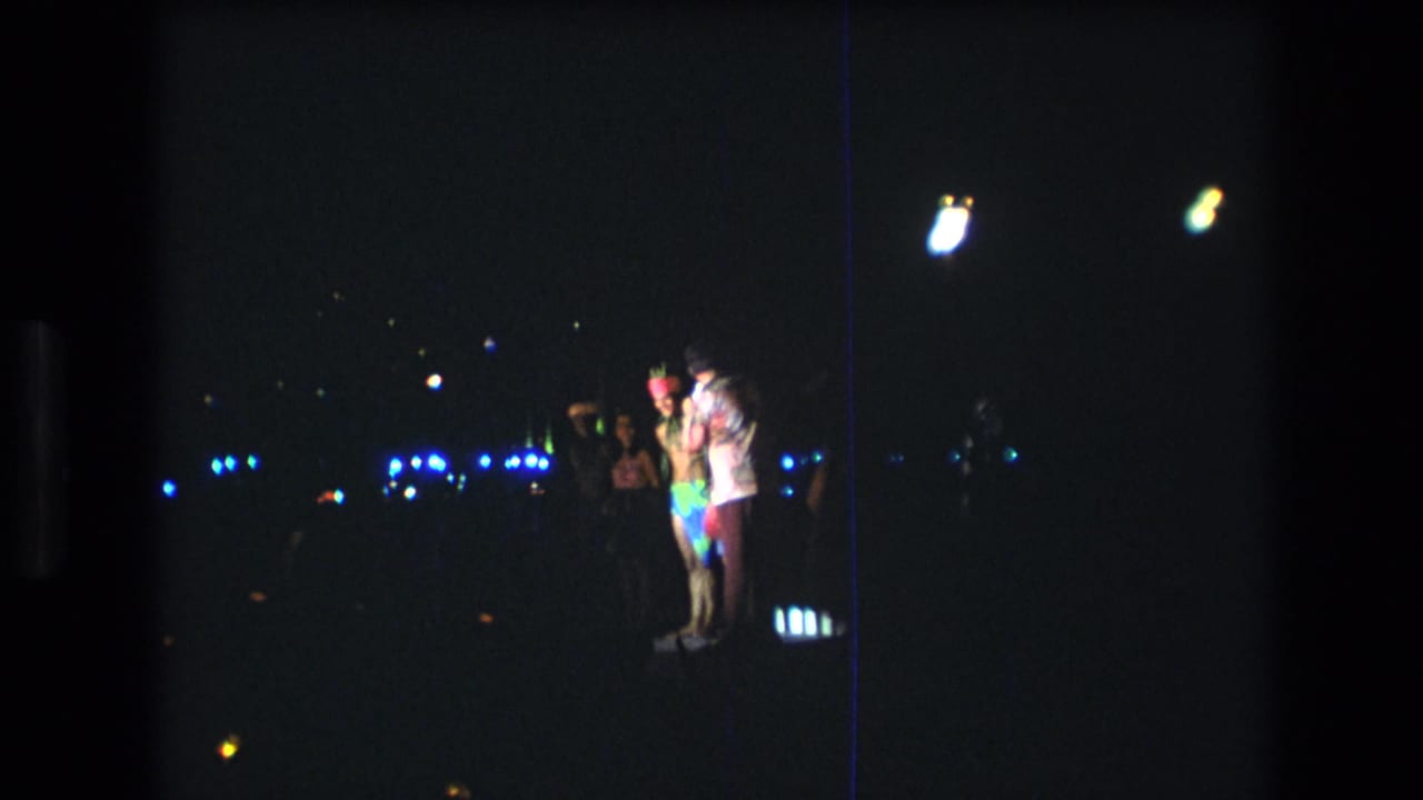 1977 Honolulu night scene with a small group standing together, including a person in bright clothing, against distant blue lights and heavy 8mm grain.