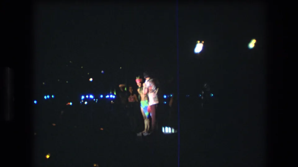 1977 Honolulu night scene with a small group standing together, including a person in bright clothing, against distant blue lights and heavy 8mm grain.