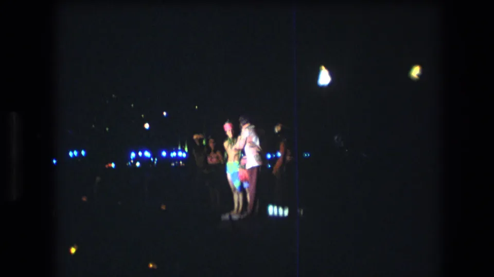 Home-movie frame from 1977 Honolulu showing a brightly dressed person beside others at a nighttime outdoor event, with blue lights and film flares in the background.