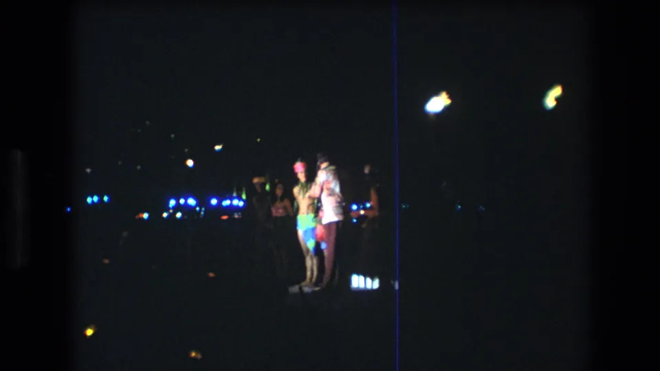 1977 Honolulu 8mm frame with people standing at a nighttime gathering, surrounded by distant blue lights, white flares, and deep shadow.