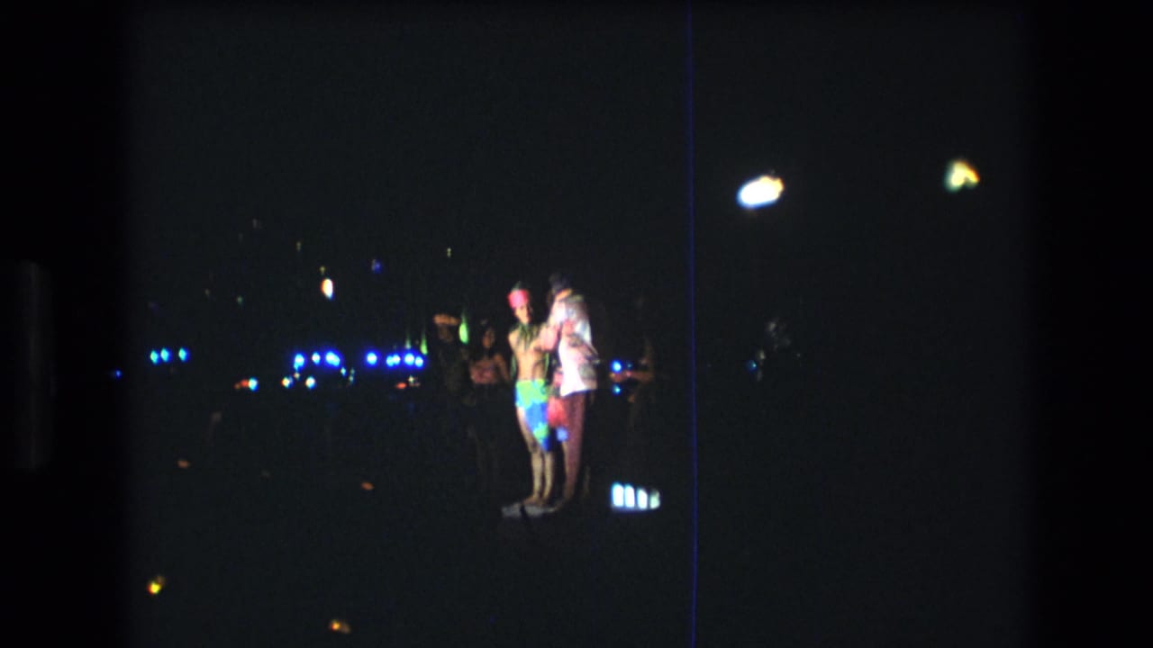 1977 Honolulu home-movie frame showing several people in bright clothing gathered and dancing under scattered blue and yellow lights at night.