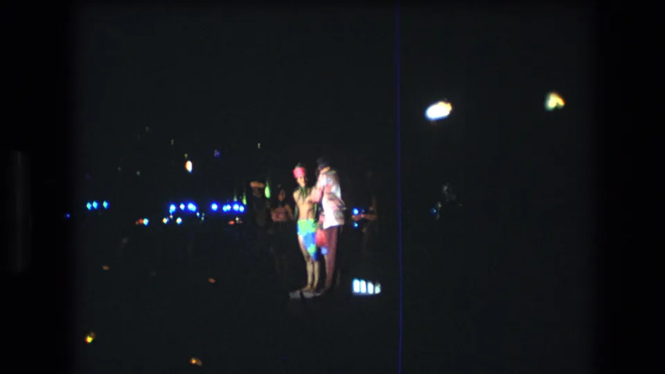 1977 Honolulu home-movie frame showing several people in bright clothing gathered and dancing under scattered blue and yellow lights at night.