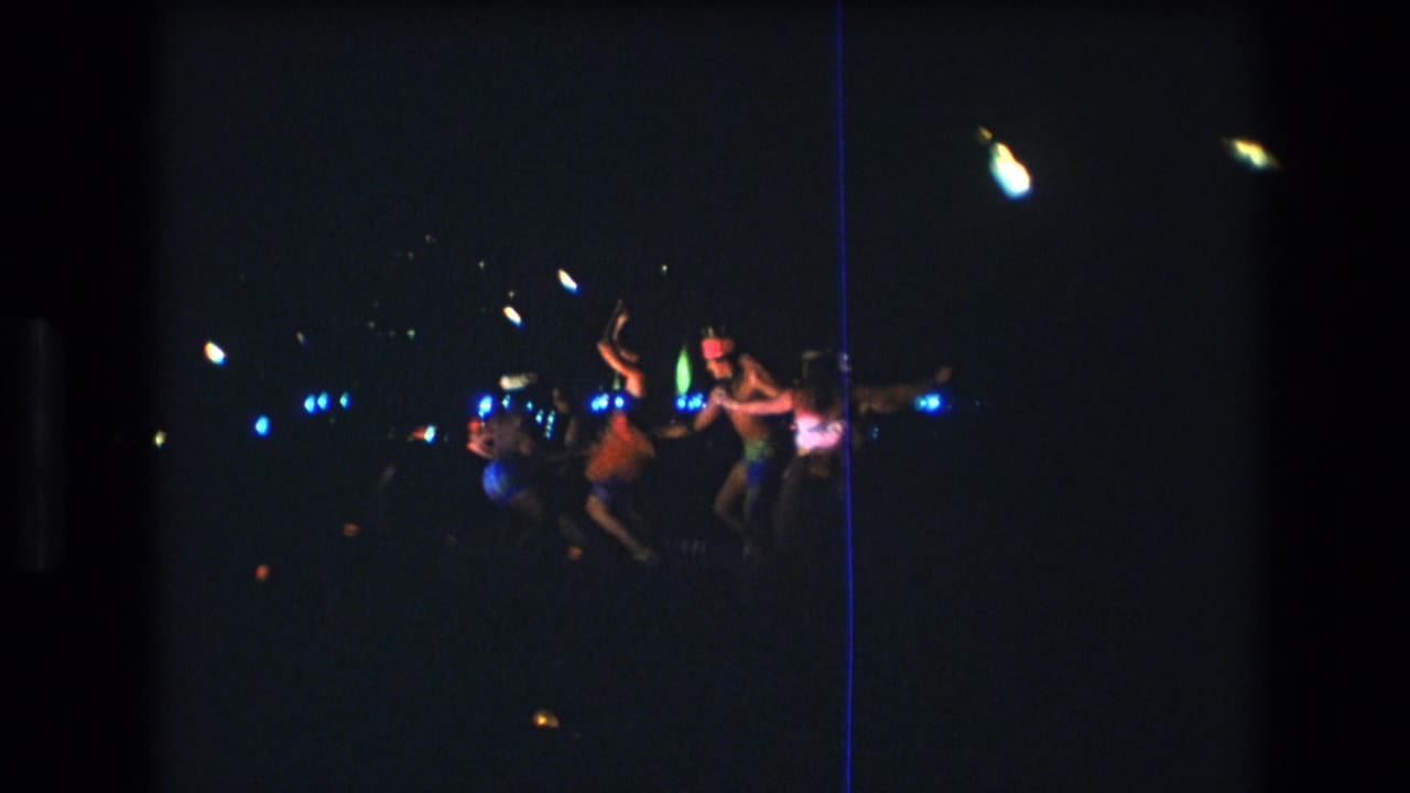 1977 Honolulu 8mm frame showing distant luau dancers performing at night under blue and white lights in a dark outdoor setting.
