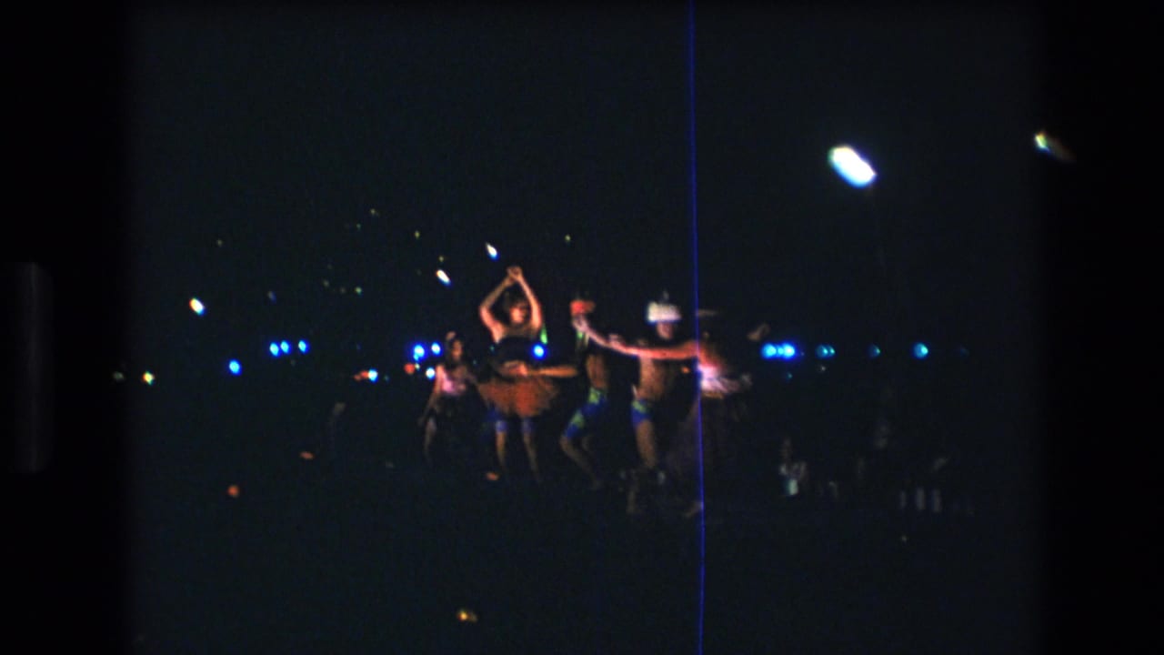 A group of dancers moves together beneath bright overhead lights at night.
