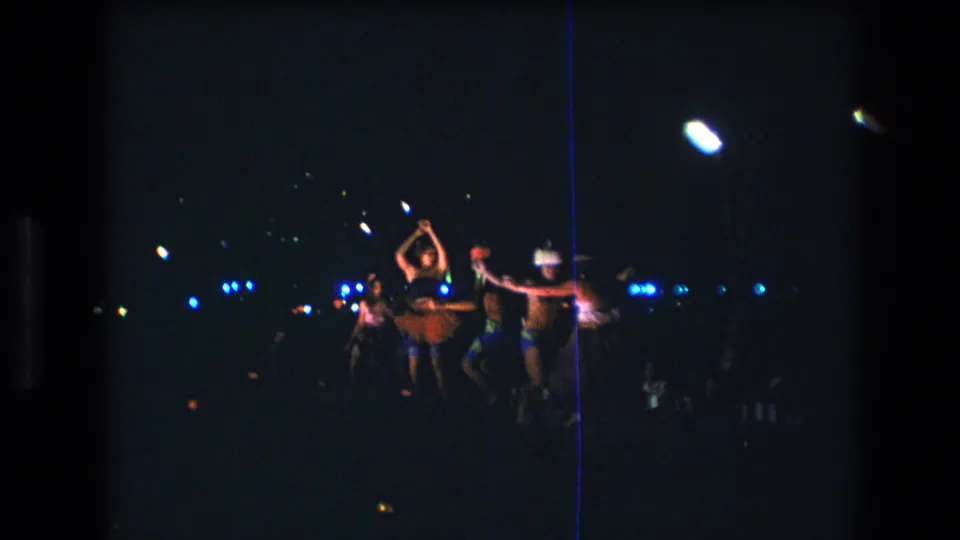1977 Honolulu 8mm frame with a small group of luau dancers moving together beneath bright nighttime lights against a dark background.