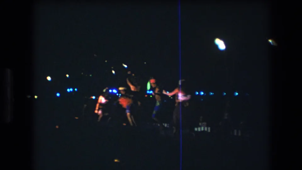 1977 Honolulu 8mm frame showing distant dancers in motion during a nighttime luau performance with blue lights and heavy film grain.