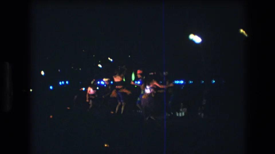 1977 Honolulu 8mm frame showing several luau dancers from a distance, with blue lights glowing across the dark nighttime performance area.