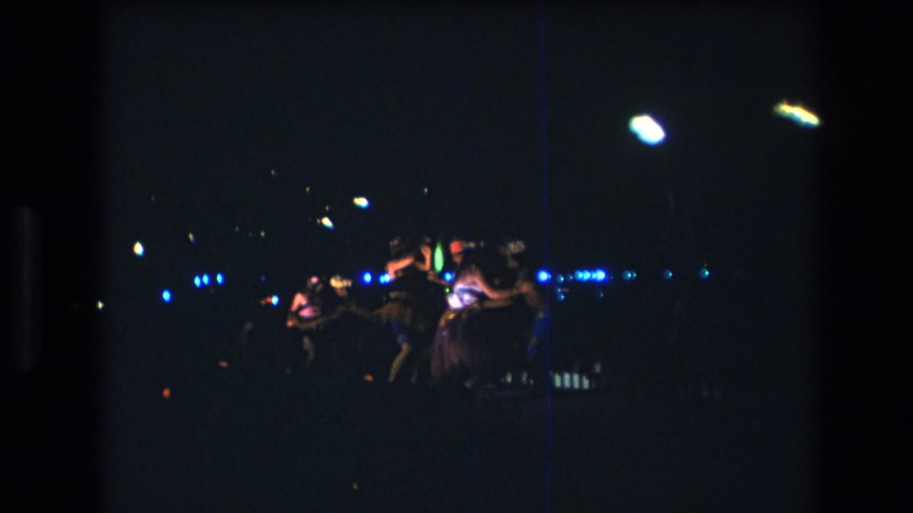 1977 8mm home-movie frame from Honolulu showing Hawaiian dancers at a distance on a dark stage with blue and white performance lights.