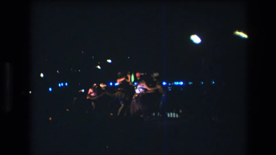 1977 8mm home-movie frame from Honolulu showing Hawaiian dancers at a distance on a dark stage with blue and white performance lights.