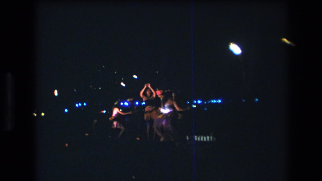 Stage dancers continue performing beneath bright lamps and blue background lights.