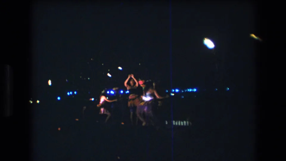 Distant 1977 Honolulu stage-performance frame with dancers moving under bright white lamps and rows of blue lights in a dark venue.
