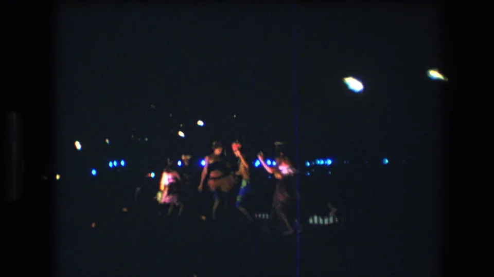1977 8mm Honolulu home-movie frame showing several Hawaiian dancers grouped onstage, filmed from a dark audience viewpoint with colored lights behind them.