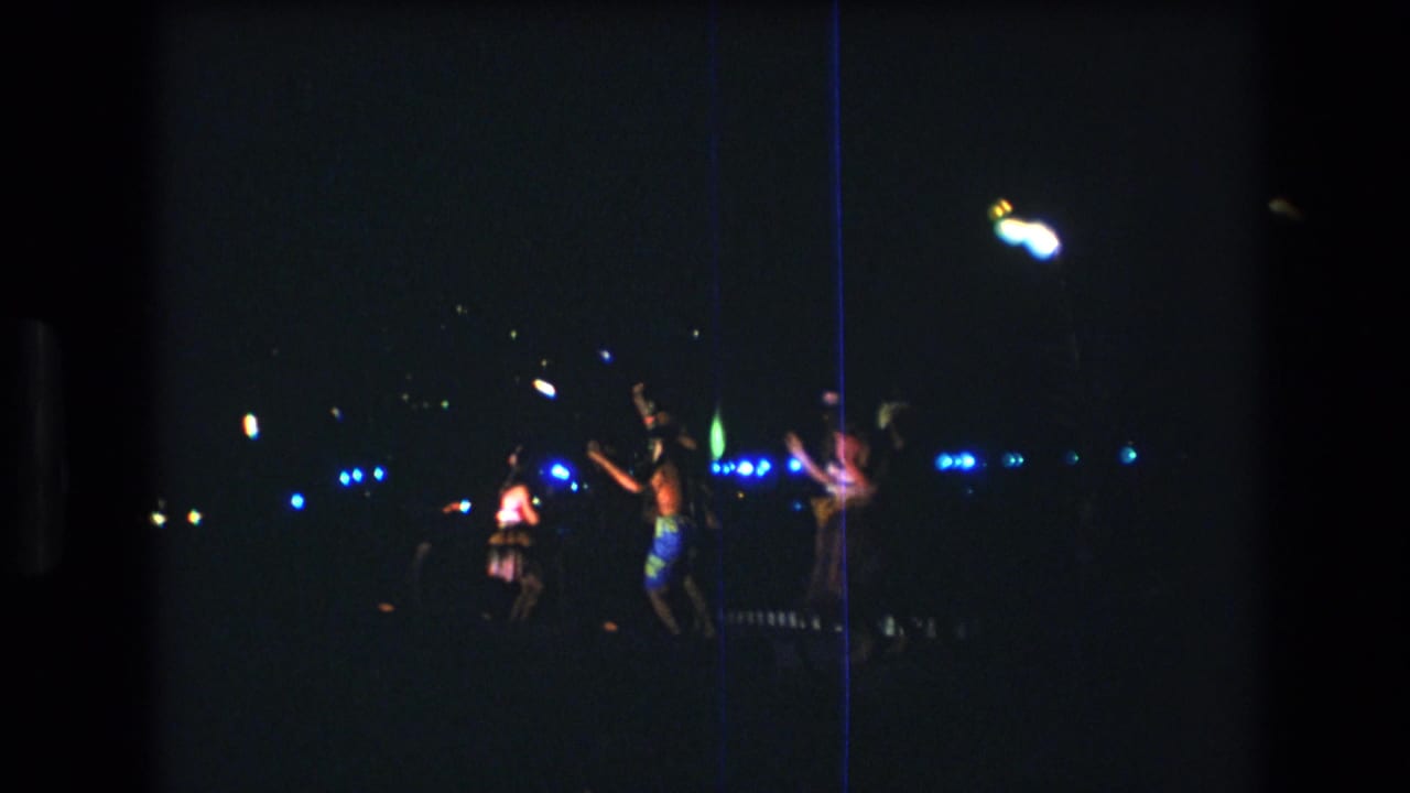 Dancers move across a dark outdoor stage with bright lights behind them.