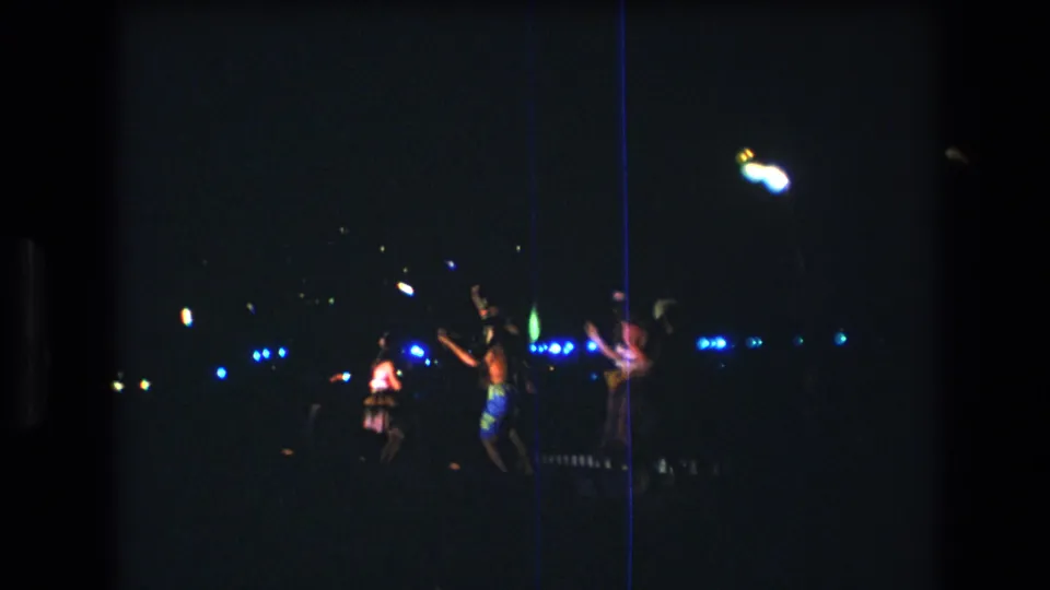 Women in colorful clothing appear mid-dance during a 1977 Honolulu night show, filmed from a distance with strong blue and white light points behind them.