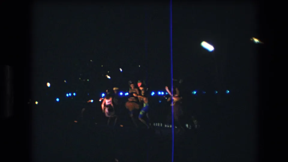 A small group of women appears in motion during the Honolulu night performance, with underexposed figures, blue lights, and visible 8mm film marks.