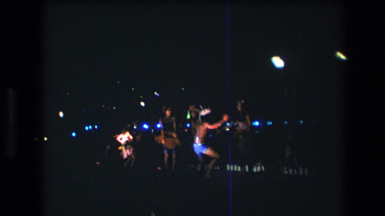 1977 Honolulu Oahu 8mm frame showing adults dancing outdoors at night, with blue background lights, colorful clothing, and a bright flame-like light source.