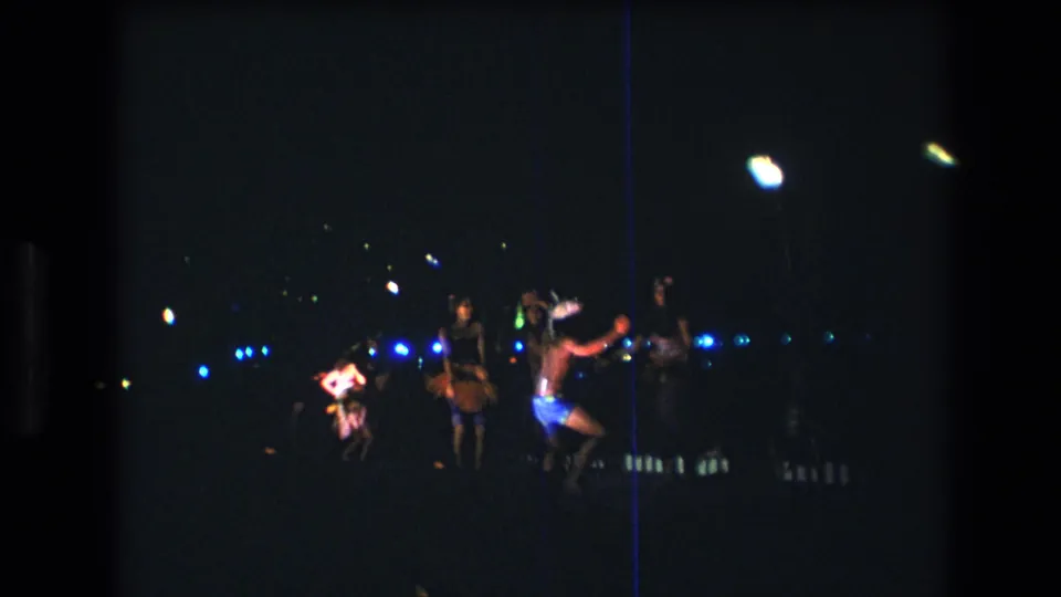 1977 Honolulu Oahu 8mm frame showing adults dancing outdoors at night, with blue background lights, colorful clothing, and a bright flame-like light source.