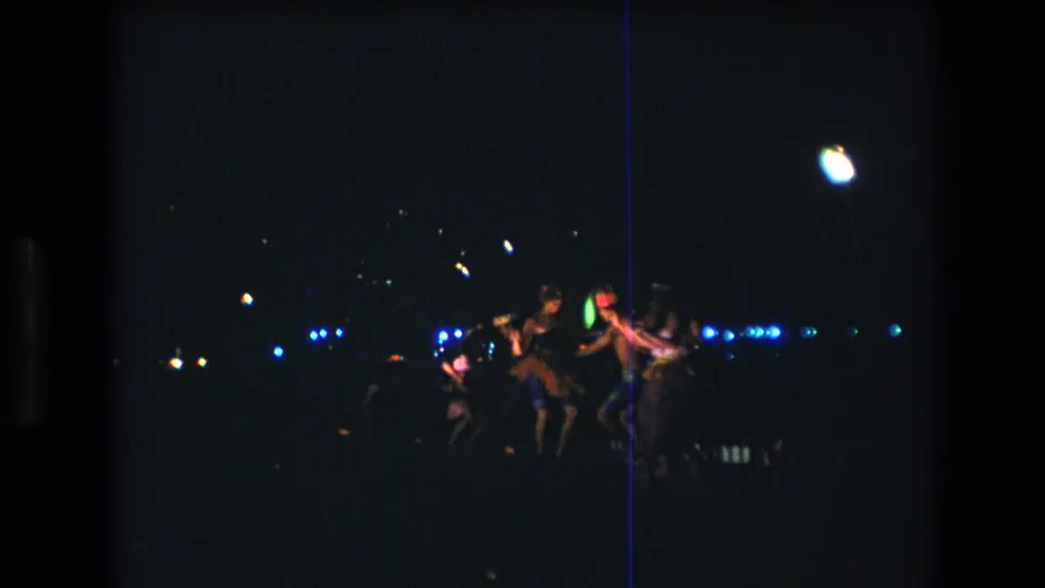 1977 Honolulu Oahu 8mm frame of costumed adults dancing together at night, partly silhouetted by scattered blue lights and a bright overhead highlight.