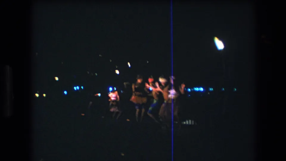 1977 Honolulu Oahu 8mm frame showing adults dancing in low light, with motion blur, colorful clothing, blue background lights, and a bright flame-like source near the group.