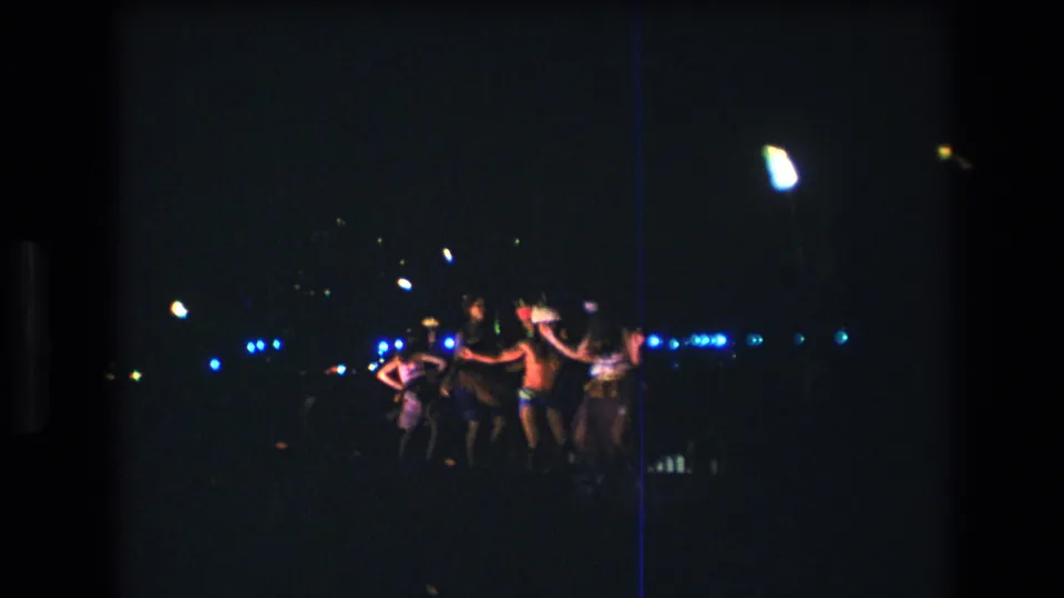 1977 Honolulu Oahu 8mm frame showing adults dancing in a clustered line at night, with blue lights across the background and strong low-light film grain.