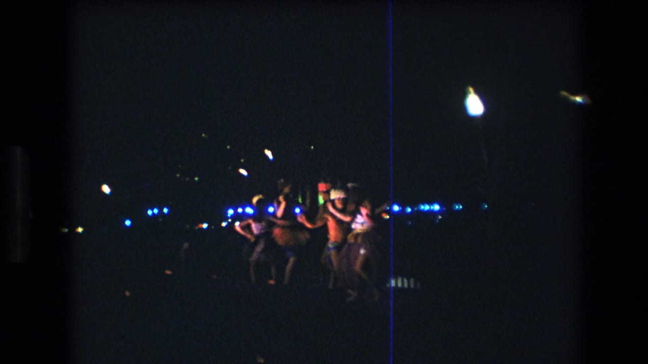 In supported 1977 Honolulu home-movie footage, several young adults dance together at a dark outdoor beach party with distant blue and white lights behind them.