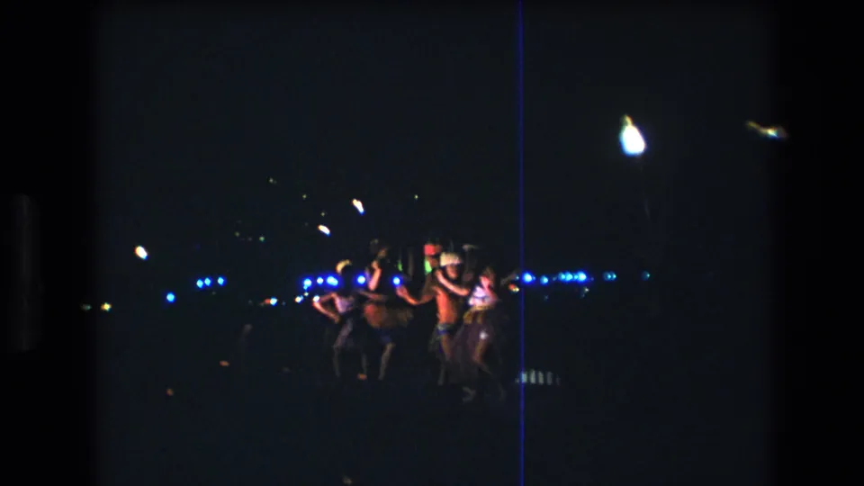 In supported 1977 Honolulu home-movie footage, several young adults dance together at a dark outdoor beach party with distant blue and white lights behind them.