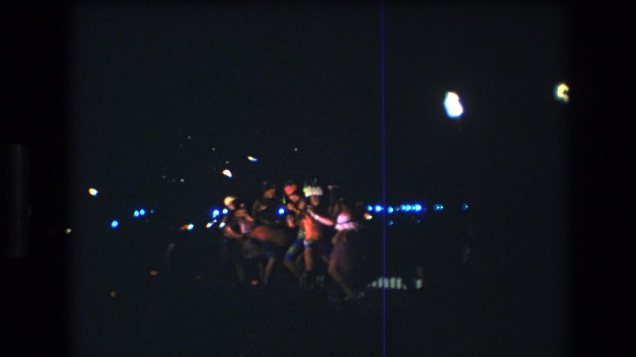 Dancers cluster together in a dark outdoor scene with blue lights in the background.