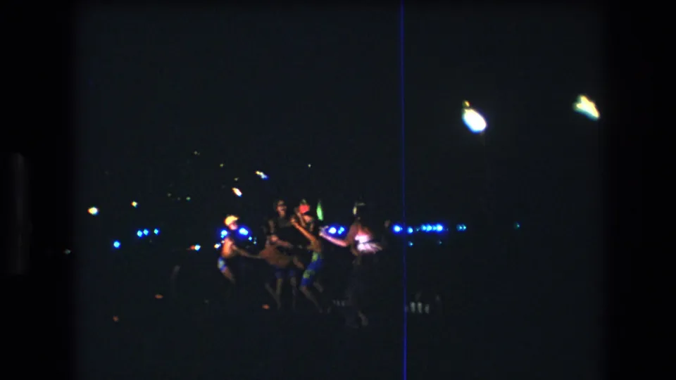 A small group of friends continues dancing in the dark beach-party scene, with the 8mm frame showing underexposed color, scattered lights, and a visible film streak.