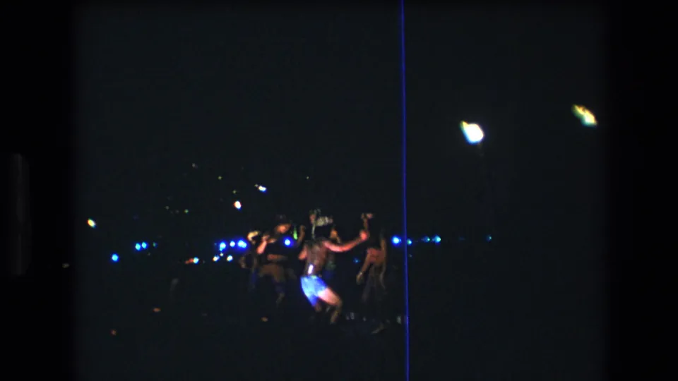 1977 8mm home-movie frame from Oahu showing a small group of luau dancers at night, seen from a distance in very low light.
