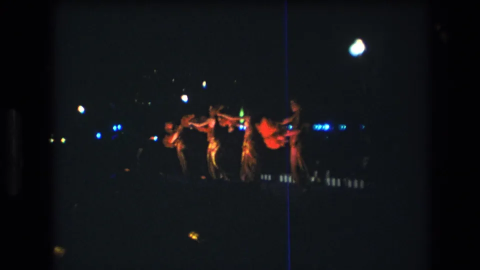 Oahu luau dancers appear in a loose line at night in this 1977 8mm frame, with deep shadow, colored lights, and visible film grain.