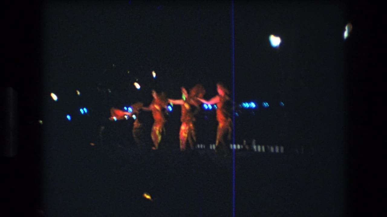 Adult dancers in flowing skirts perform on a distant outdoor stage at night in Honolulu, consistent with the supplied 1977 home-movie metadata.