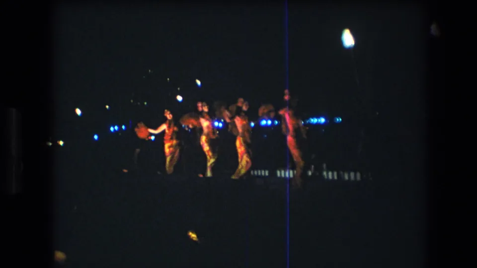 A group of adult Hawaiian dancers appears in formation on a nighttime outdoor stage in Honolulu, with red and blue lighting visible in the dark scene.