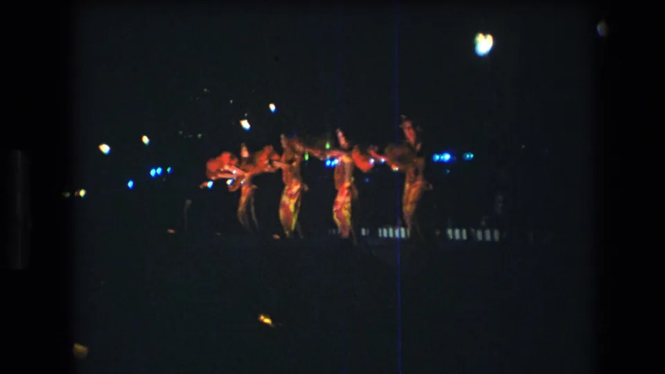 Adult dancers continue a synchronized outdoor stage routine at night, seen from a distant audience viewpoint in the 1977 Honolulu home-movie clip.
