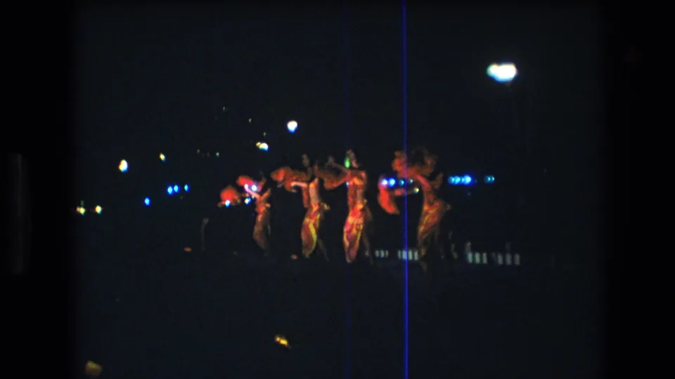 The closing frame shows adult dancers on the outdoor stage, partly silhouetted by bright lamps and colored lights in the Honolulu nighttime performance.
