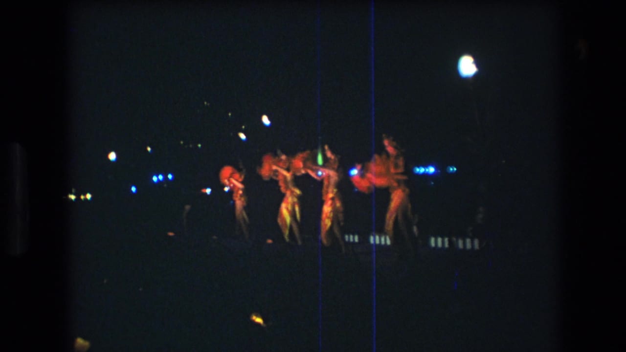 Four dancers in red sparkly leotards appear on a dim Honolulu stage in 1977 home-movie footage, with blue points of light and a bright overhead lamp behind them.
