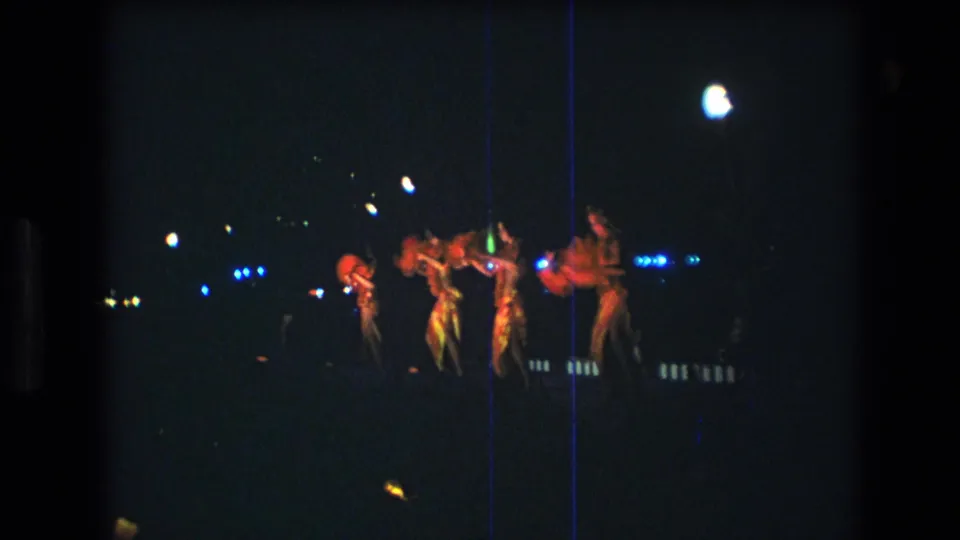 Four dancers in red sparkly leotards appear on a dim Honolulu stage in 1977 home-movie footage, with blue points of light and a bright overhead lamp behind them.
