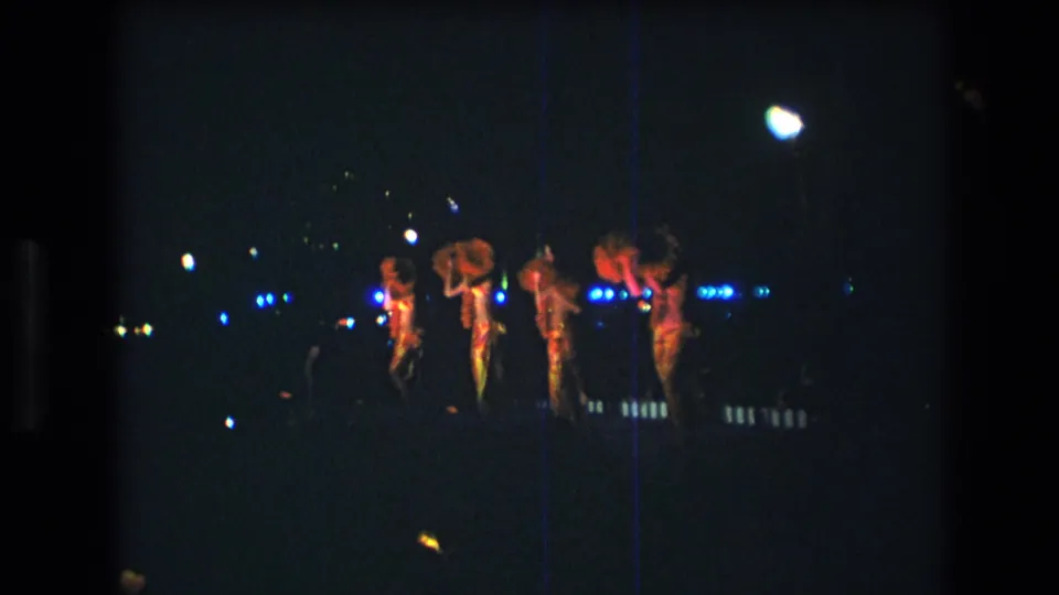 The group continues synchronized choreography on a low-lit Honolulu stage, with red costumes catching the light and film scratches visible across the frame.