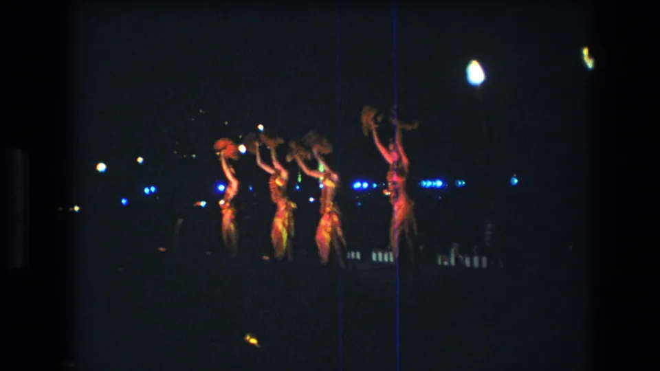 Four performers in red stage costumes raise their arms in unison during the 1977 Honolulu performance, framed from a distant audience perspective on grainy 8mm film.