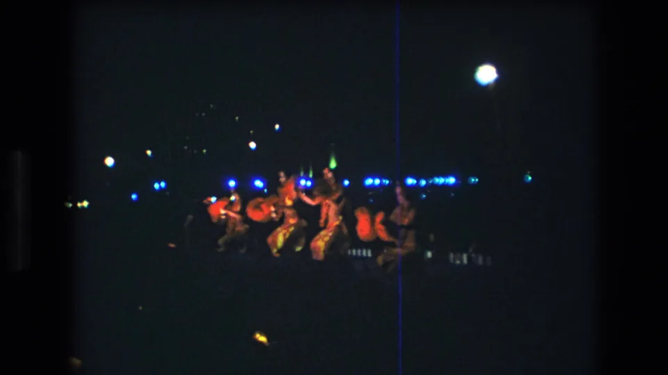 The performers move into a lower clustered pose on the dim Honolulu stage, with red costumes, blue stage lights, and pronounced Super 8 grain visible.