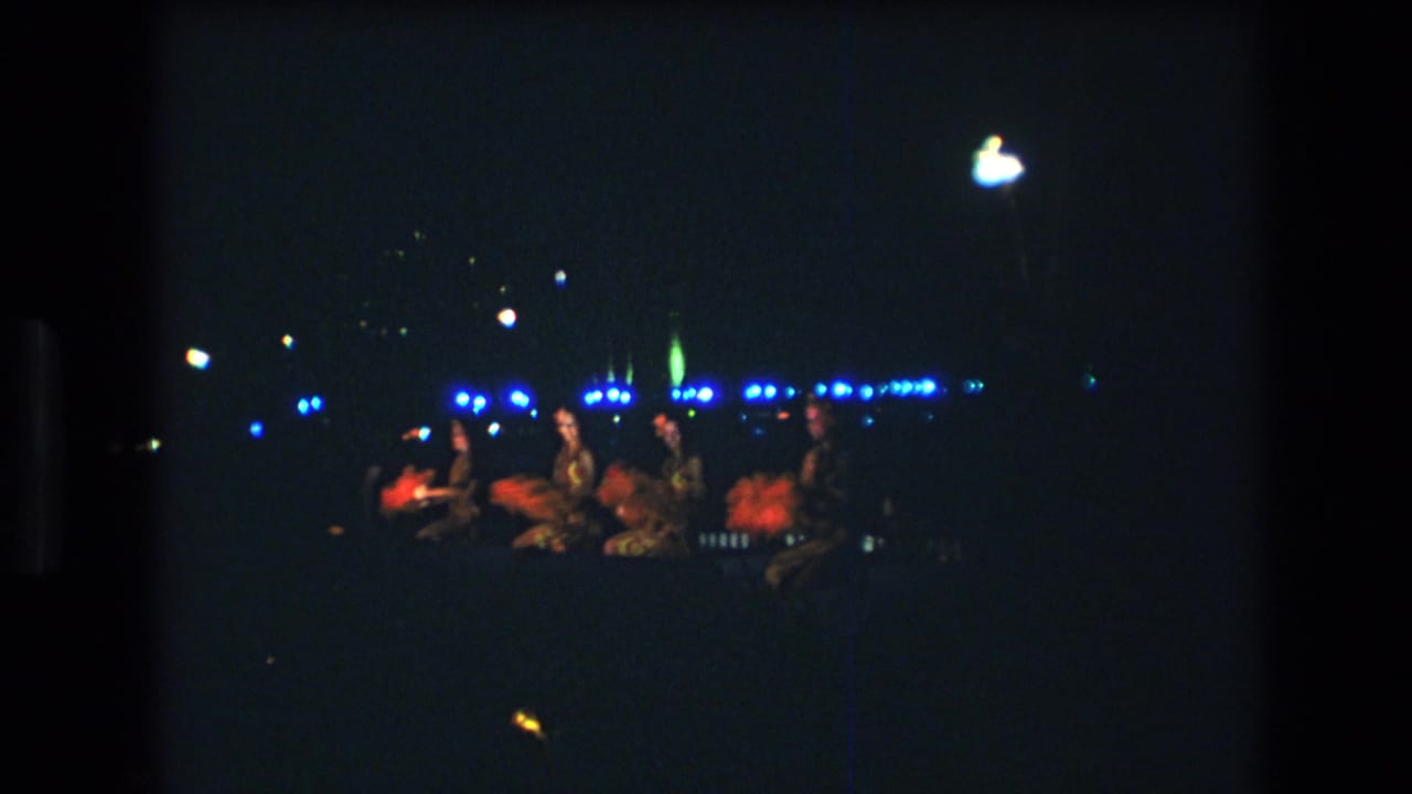 1977 Oahu home-movie frame showing distant performers on a dimly lit luau stage with blue lights and red-orange costume accents.