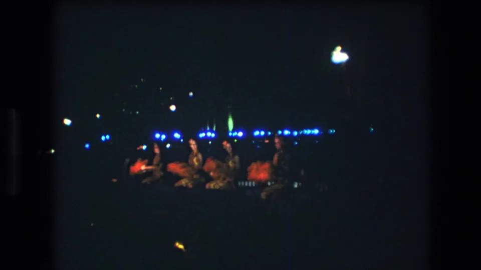 1977 Oahu home-movie frame showing distant performers on a dimly lit luau stage with blue lights and red-orange costume accents.