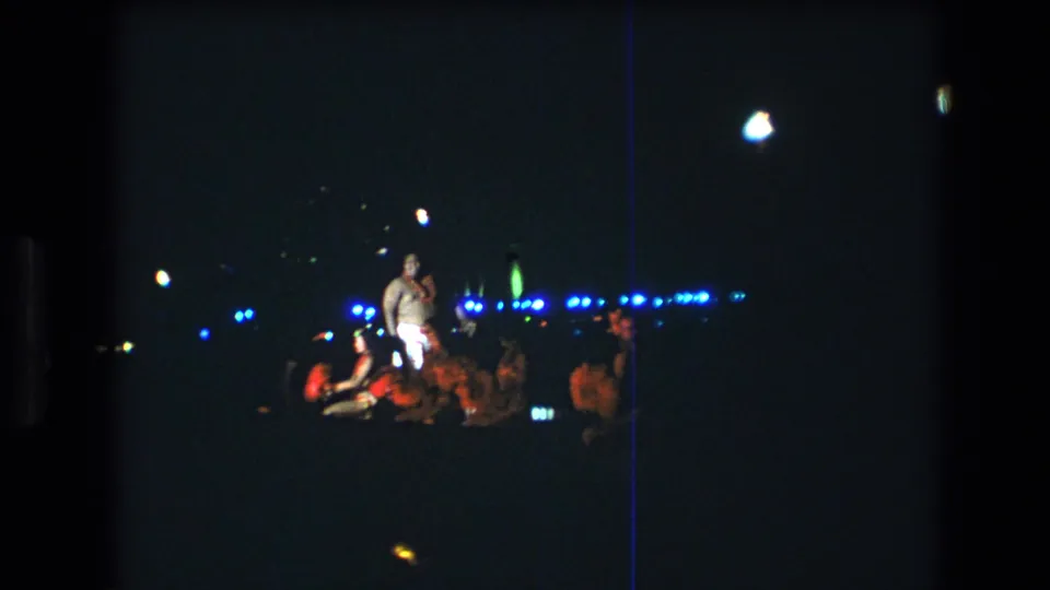 1977 Oahu luau frame with a male performer in light pants standing beside seated dancers on a dark, distant stage.