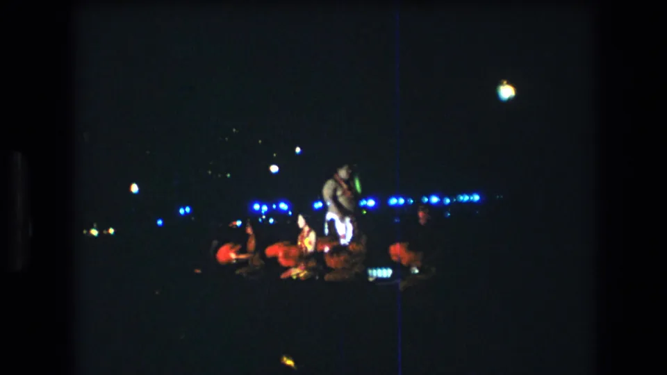 Distant home-movie view of a 1977 Honolulu Oahu luau stage with a male performer and seated dancers under blue lights.