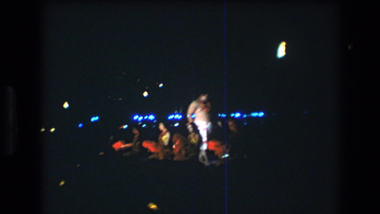 In 1977 Honolulu, a shirtless lei-wearing performer appears on a dim night stage while a seated audience watches under blue background lights.