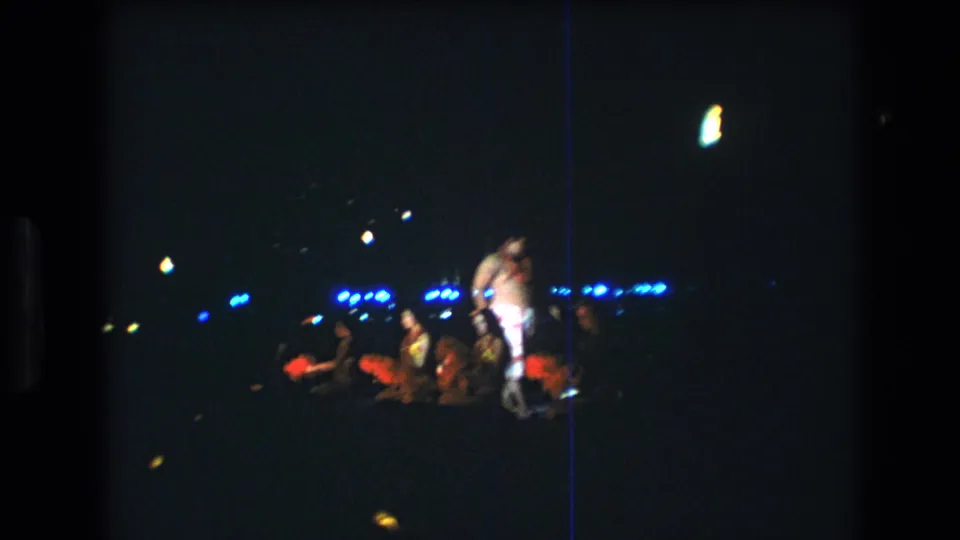In 1977 Honolulu, a shirtless lei-wearing performer appears on a dim night stage while a seated audience watches under blue background lights.