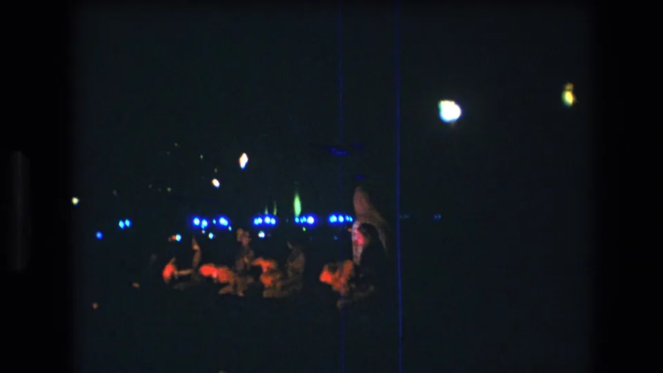 The Honolulu night performance continues from a distance, with spectators clustered in the foreground and blue stage lights glowing behind the performer area.